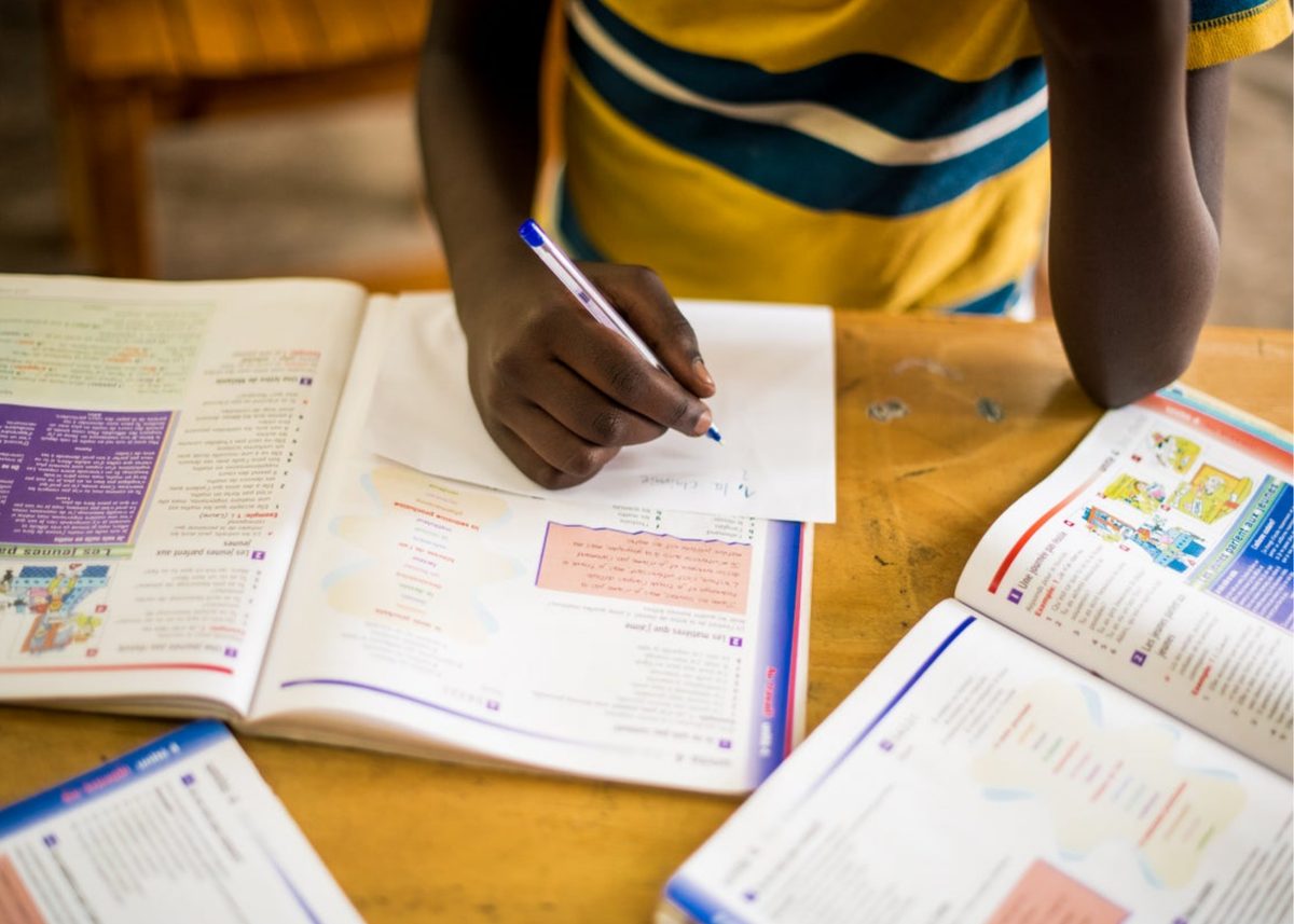 A student writing at a desk with open workbooks spread across the table